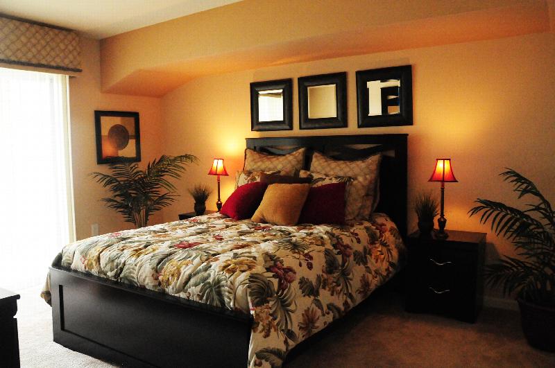 Village at Stratford for ages 55 and above shows a queen sized bed with two end tables. There is wall to wall carpeting. several plants and pieces of wall art decorate the room.