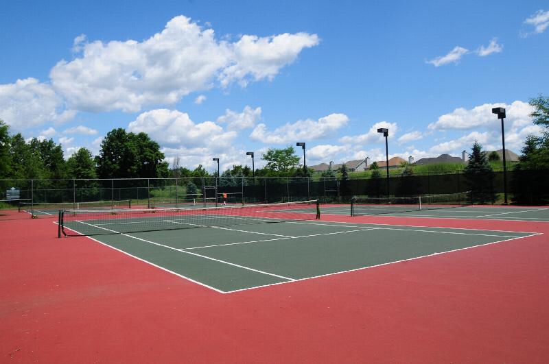 Pike Run Affordable Tennis Court photo showing several tennis courts surrounded by a fence. There are lampposts that provide lighting to the tennis courts. There are several bushes and trees that highlight the landscape. The apartments can be seen in the background.