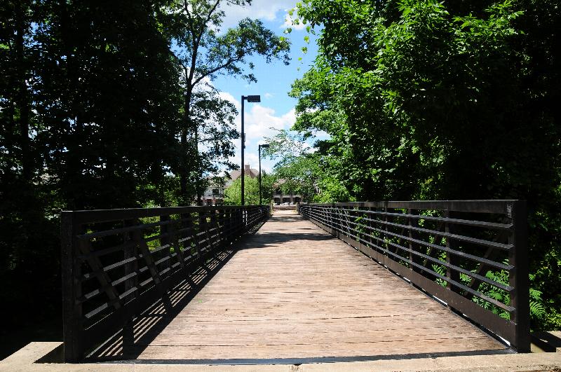Pike Run Affordable exterior photo showing a wooden bridge and several lampposts. The apartments can be seen in the background. Several bushes and trees highlight the landscape.