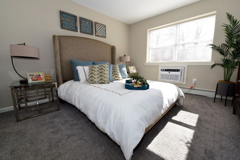 Photo of a bedroom in a refurbished apartment at Edison Village plush wall to wall carpeting, ned with 2 end tables and lamps, and a large double window to allow for natural light
