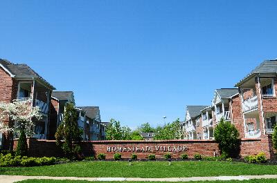 Homestead Village exterior photo of the property featuring a wall with the Homestead Village name. There are bushes and green grass landscaping the area. Apartment buildings can be seen to both the left and the right of the wall.