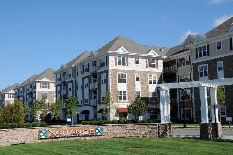 Helix at Xchange Affordable exterior photo shows a four story apartment building with stone and beige colored siding, red shutters and white trim. Photo also shows partial pergola, community logo signage fixed to a stone facade surrounded by green grass and tree lined streets.