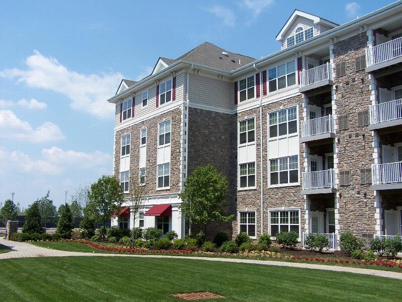 Helix at Xchange Affordable exterior photo shows a four story apartment building with stone and beige colored siding, red shutters and white trim. Landscaped court yard can be seen with garden style shrubs, trees, paved walkway and park benches for seating.