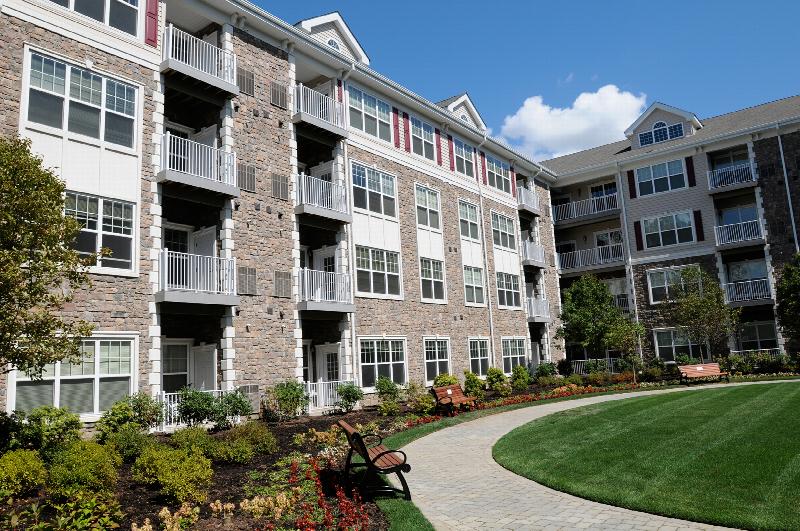 Helix at Xchange Affordable exterior photo shows a four story apartment building with stone and beige colored siding, red shutters and white trim. Landscaped court yard can be seen with garden style shrubs, trees, paved walkway and park benches for seating.