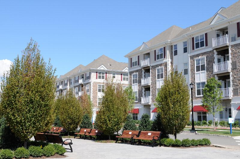 Helix at Xchange Affordable exterior photo shows tree lined community courtyard with paved walkway and several park benches for seating. Four story apartment building with stone and beige colored siding, red shutters and white trim can be seen the background.
