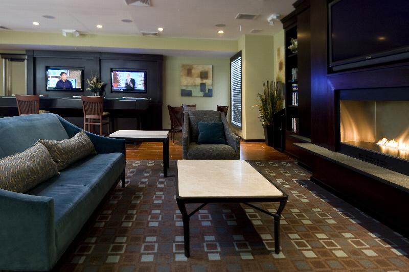 Helix at Xchange Affordable photo of amenity room showing a couch and coffee table in front of a fireplace with a TV display located above it. A large bookcase, additional seating and computer work stations can be seen in the background.   