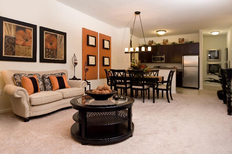 Helix at Xchange Affordable interior photo of living room shows couch with decorative pillows, round coffee table and two side tables. Adjacent to the living room is a dining table that seats up to six with a flower center piece.  The room is carpeted with the exception of the kitchen area seen in the background. The kitchen has expresso cabinets, stainless steel appliances and ceramic tiled flooring.