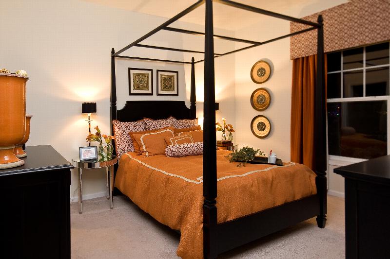 Helix at Xchange Affordable interior photo of master bedroom shows a four post king sized bed with decorative pillows and two night tables located on either side. Two large dressers can be seen in the foreground. 