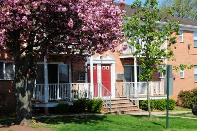 Woodbridge Gardens exterior photo shpws a brick apartment building with red front doors and white trim. Stairs lead to the apartment entry and there are balconies on both the first and second floors. Green grass, shrubs and trees landscape the surrounding area.