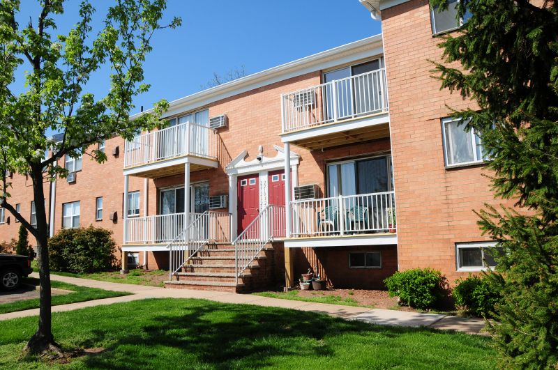 Woodbridge Gardens exterior photo shpws a brick apartment building with red front doors and white trim. Stairs lead to the apartment entry and there are balconies on both the first and second floors. Green grass, shrubs and trees landscape the surrounding area.