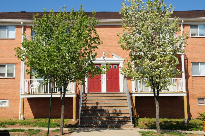 Woodbridge Gardens photo features 2 trees on either side of a sidewalk that leads to stairs heading up to the apartment entry. The building is brick with red doors and white trim. Balconies can be seen on both the first and second floors.