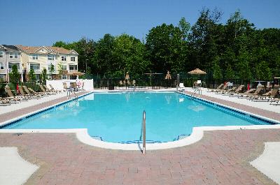 Princeton Terrace swimming pool photo showing a very large pool with reclining chairs located on both sides surrounded by a fence. The apartment building can be seen in the background. Several trees highlight the landscaping.