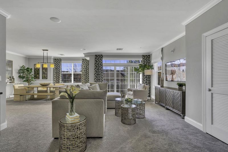 Interior Photo of the model at Princeton Terrace at West Windsor showing a living room with seating area and wall to wall carpeting. Sliding glass doors that lead to the balcony. The dining area with table can be seen in the background