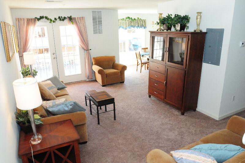 Harbortown Heights interior photo of the living room showing wall to wall carpeting, a couch and chair, end tables, a coffee table and an entertainment center. French doors lead to a balcony. Eat in dining room area is shown in the background.