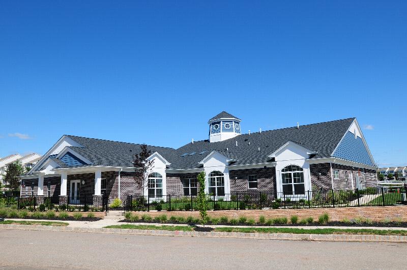Exterior image of the Harbortown Clubhouse showing a large brick building with a covered entrance. There is a black metal fence surrounding the building, and many bushes, plants, flowers and trees landscape the outside.