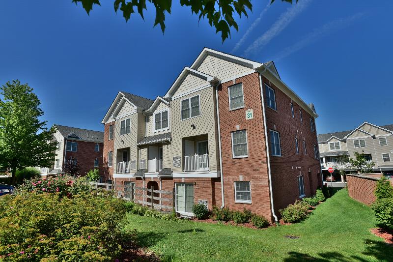 Exterior photo of the Village at Sun Valley showing a red brick apartment building with tan siding. There are balconies on the second floor, and a large grassy area surround the building, Several trees and many bushes landscape the area.
