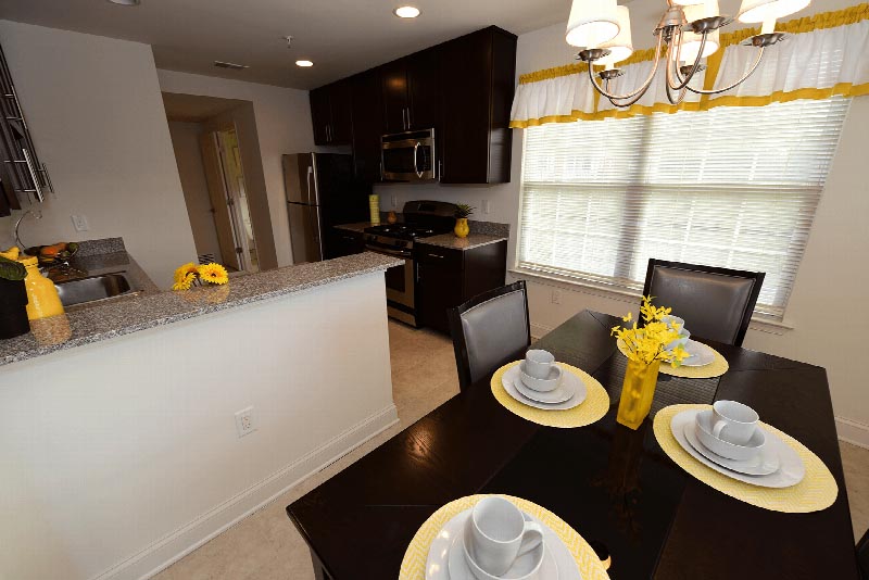 Photo of a dining area and kitchen at the Village at Sun Valley. There are dark kitchen cabinets, granite countertops, stainless steel appliances in the kitchen and a table for four with table settings in the dining area. There is a large double window to allow for natural light.