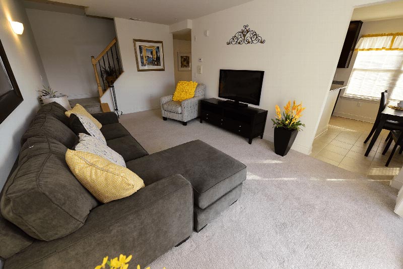 Village at Sun Valley photo of a living room showing a sectional couch, a chair, an entertainment center with a TV. There are several plants and wall decorations. Plush wall to wall carpeting fills the room and leads to the stairway to another level.