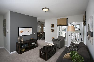 Autumn Hills photo of the living room showing a couch and chair, a coffee table and media console. There is a TV on the wall. There are pillows on the couch and other decorations throughout the room. A door to the balcony and a window letting in natural light are also shown.