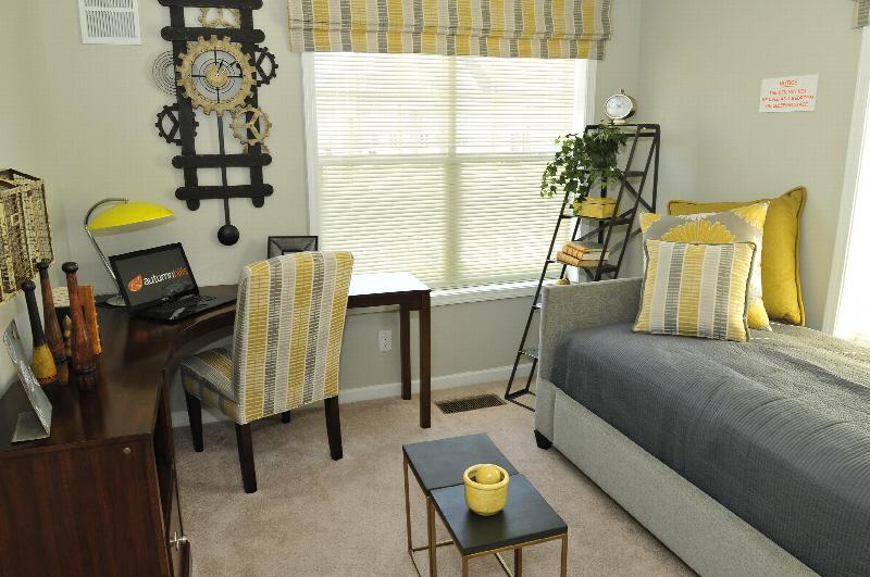 Autumn Hills photo of the den shows a corner desk with a laptop computer, a daybed along the wall. There are window blinds and a large wall clock. Throw pillows and other decorations fill the room.