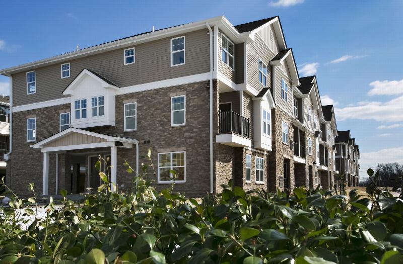 Autumn Hills exterior photo shows a side entrance, a combination of both stone and siding, with white trim and windows. Lush green bushes are featured in the foreground.