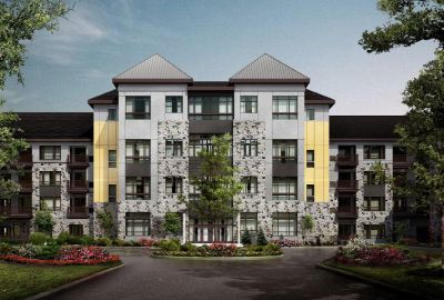 Next at Xchange exterior photo shows five story apartment building with light colored brick and stone facade. Garden style shrubs, green grass and trees highlight the surrounding landscaping