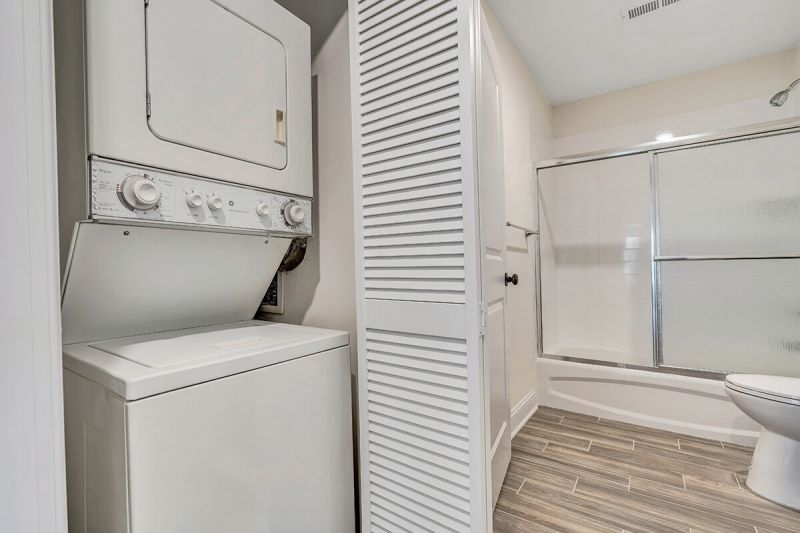 Interior photo of a renovated apartment at Pike Run Village, showing a bathroom with wood grain tile floor, new tub with shower doors, new toilet and a closet with new stackable washer and dryer.