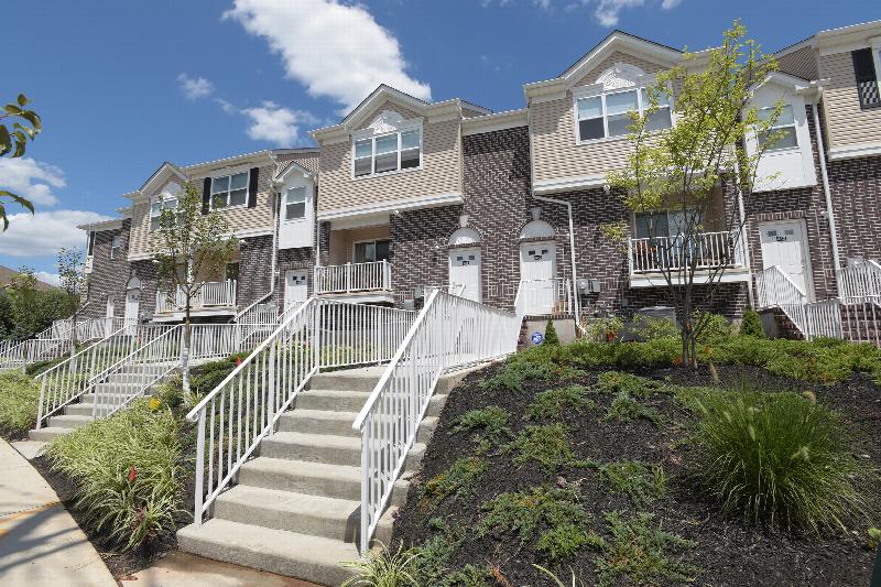 Exterior photo of a Harbortown Sail apartment building made of dark brown brick and cream siding, showing staircases leading to several different entrances. Newly planted bushes and trees landscape the area.