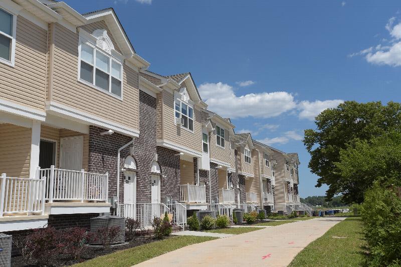 Exterior photo of a Harbortown Sail apartment building made of dark brown brick and cream siding, showing staircases leading to several different entrances. There are first floor balconies and newly planted bushes and trees landscape the area.