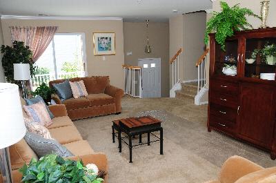 Interior photo of the living room at Harbortown Sail. Wall to wall carpeting fills the room. There is a couch and loveseat with two end tables and a coffee table. Stairs leading down to the entranceway and stairs leading up to additional living areas can be seen.
