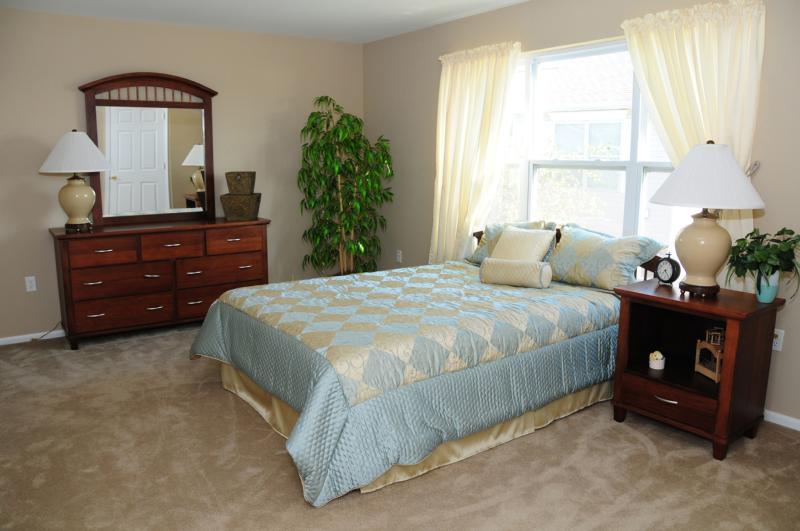 Harbortown Sail bedroom photo showing a queen sized bed with an end table and dresser. The room features wall to wall carpeting and large windows to allow for natural light.