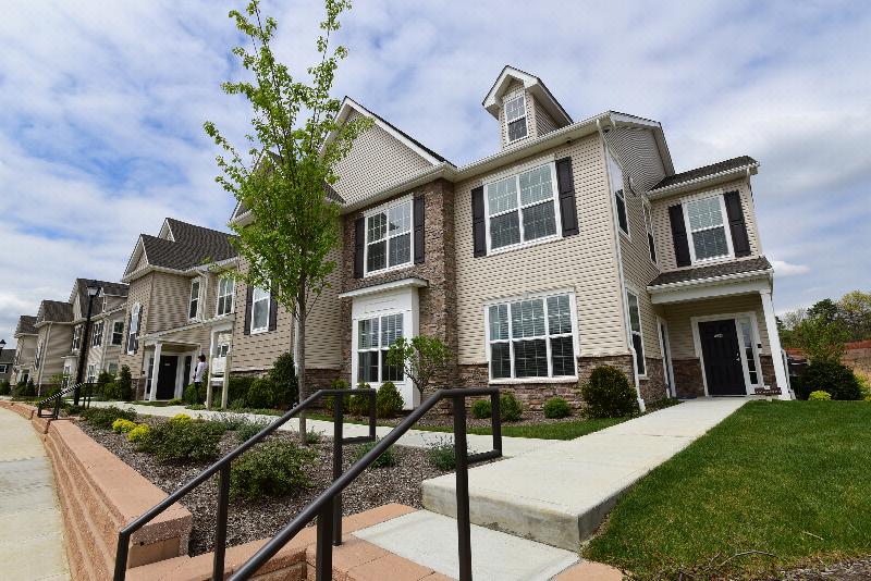 Woodhaven Commons exterior photo shows a two level apartment building with stone and siding, white trim, black doors and shutters. Sidewalks lead to entry doors and landscape is shown around the building.