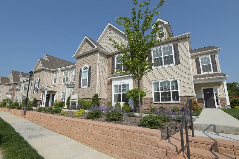Woodhaven Commons exterior photo shows a two level apartment building with stone and siding, white trim, black doors and shutters. Sidewalks lead to entry doors and landscape is shown around the building.