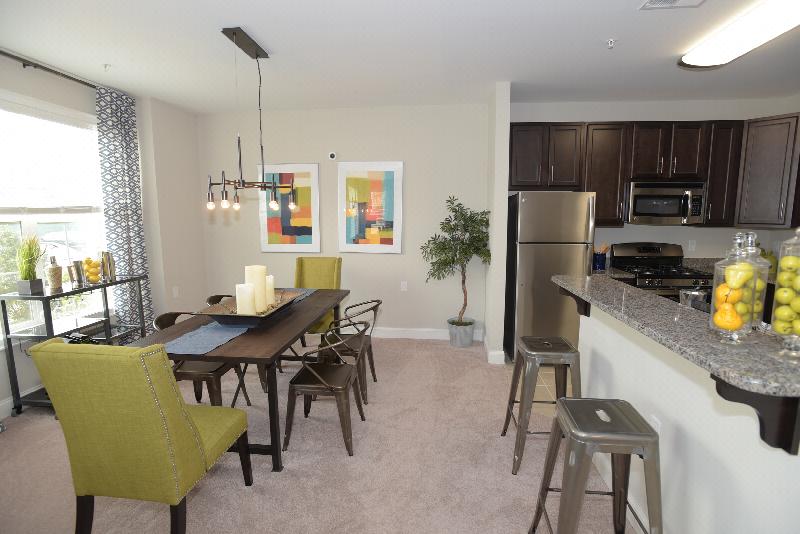 Woodhaven Commons dining room image shows a table with seating for six. Artwork is displayed on the wall, and various decorations are displayed on the table top. Part of the kitchen can be seen, with stools along the kitchen countertop.