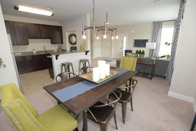 Woodhaven Commons dining room image shows a table with seating for six. Artwork is displayed on the wall, and various decorations are displayed on the table top. Part of the kitchen can be seen, with stools along the kitchen countertop. The living room can be seen in the background.