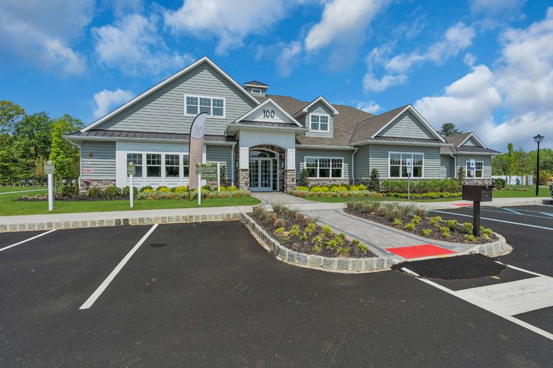 Exterior photo of the clubhouse at Woodhaven, showing a paver walkway leading to a covered double door entryway. The building is finished with mixed stone, green siding and white trim and windows