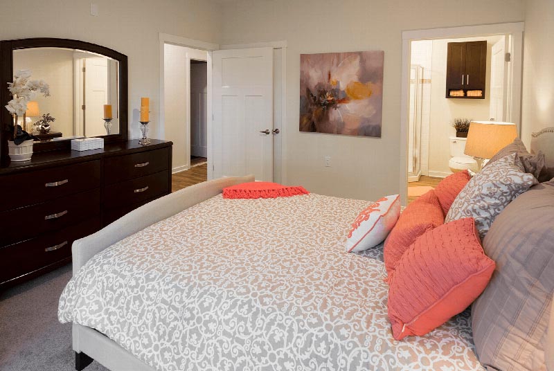 Gardens at Jackson Twenty One Interior photo showing a queen sized bed, a dresser, and a nightstand with a lamp. There are decorations on the dresser and nightstand. The bathroom can be seen in the background with a stand up shower and a toilet.