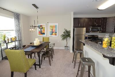 Woodhaven Commons dining room image shows a table with seating for six. Artwork is displayed on the wall, and various decorations are displayed on the table top. Part of the kitchen can be seen, with stools along the kitchen countertop.