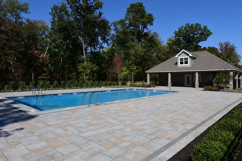 Exterior photo of the pool area at the clubhouse at Woodhaven showing a large swimming pool surrounded by a paver patio and lush landscaping with a large pool house.