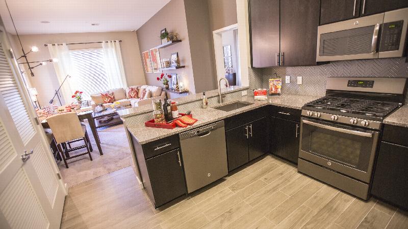 The Grove at One92 Living Room photo showing the living room from the kitchen. The kitchen has espresso cabinets, stainless steep appliances and laminate flooring. There is a sectional couch and small table in the living room. There is a 4 person dining room table and several chairs. There are decorations on the kitchen counter and the tables.