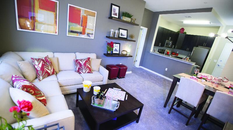 The Grove at One92 Living Room photo showing the living room. The kitchen has espresso cabinets, stainless steep appliances and laminate flooring. There is a sectional couch and small table in the living room. There is a 4 person dining room table and several chairs. There are decorations on the kitchen counter and the tables.