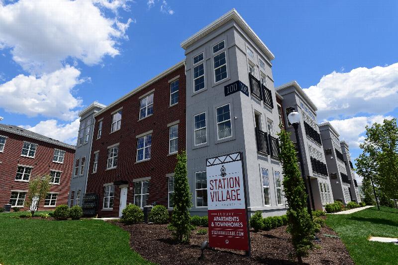 Station Village at Avenel Exterior photo shows a three story apartment building with dark reddish brick and gray stucco. Green grass and bushes landscape the area. 