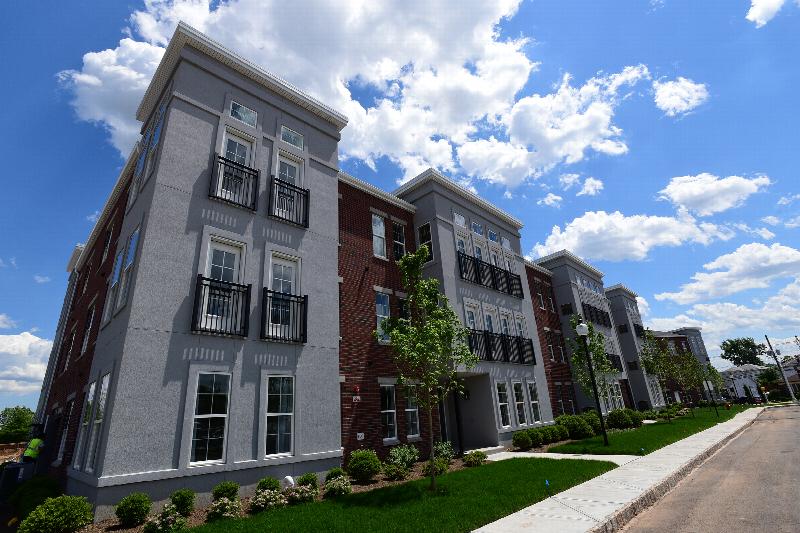 Station Village at Avenel Exterior photo shows a three story apartment building with dark reddish brick and gray stucco. Green grass and bushes landscape the area.