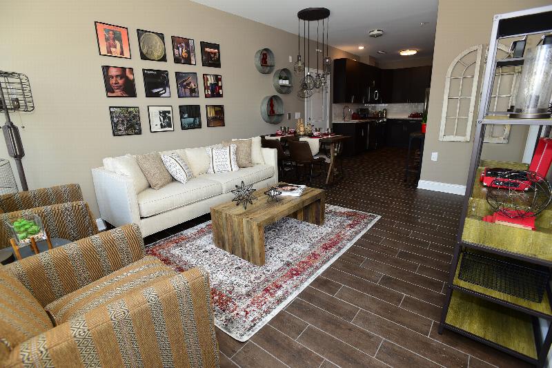 Station Village at Avenel photo of living room shows a seating area with couch, two chairs, coffee table and a media canter. Various pieces of wall art, an area rug and other items decorate the area. The dining area and kitchen are visible in the background.