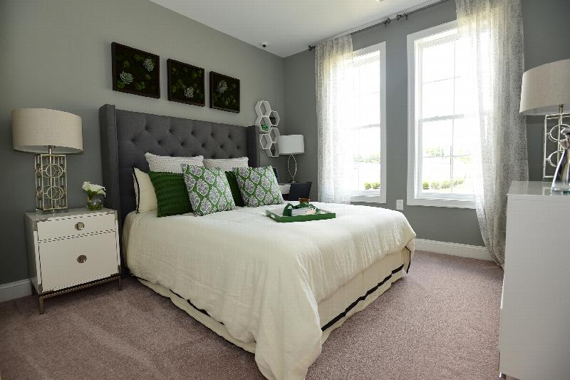 Station Village at Avenel bedroom photo shows a queen sized bed with two end tables and a dresser. There are lamps on both end tables and on the dresser. The room features two large windows to allow for natural light and lush wall to wall carpeting.