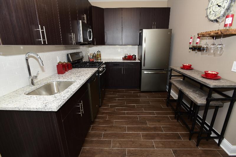 Station Village Affordable Kitchen photo showing espresso cabinets, granite counter tops and stainless steel appliances. Laminate flooring runs throughout the apartment. There is a small table and two chairs against the wall. There are decorations on the counter top, table and on the walls.