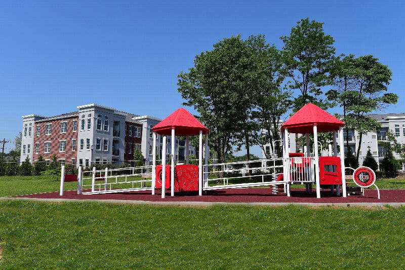 Station Village Affordable Eric Lagrand Park photo showing a playground surrounded by a fence. The apartment buildings can be seen in the background. Several bushes and trees highlight the landscape.